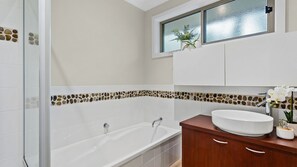 Une salle de bain avec une baignoire, un lavabo blanc, des meubles en bois et une fenêtre agrémentée d’une plante.