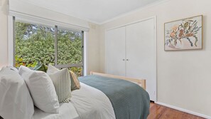 Ein Schlafzimmer mit Bett, Kissen, einem Fenster mit Blick auf Bäume, einem Kleiderschrank und einem gerahmten Bild an der Wand.