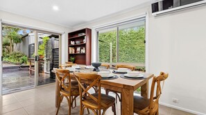 A dining area with a table set for four, wooden chairs, a bookshelf, and a view of greenery outside. - Saltwater Sanctuary | Fireplace & Walk to Beach (Mount Martha)