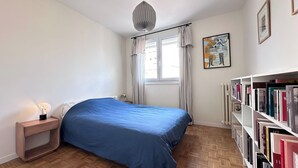 Ein Schlafzimmer mit einem Bett, einer Nachttischlampe, einem Bücherregal, einem Fenster mit Vorhängen und einer Deckenleuchte.