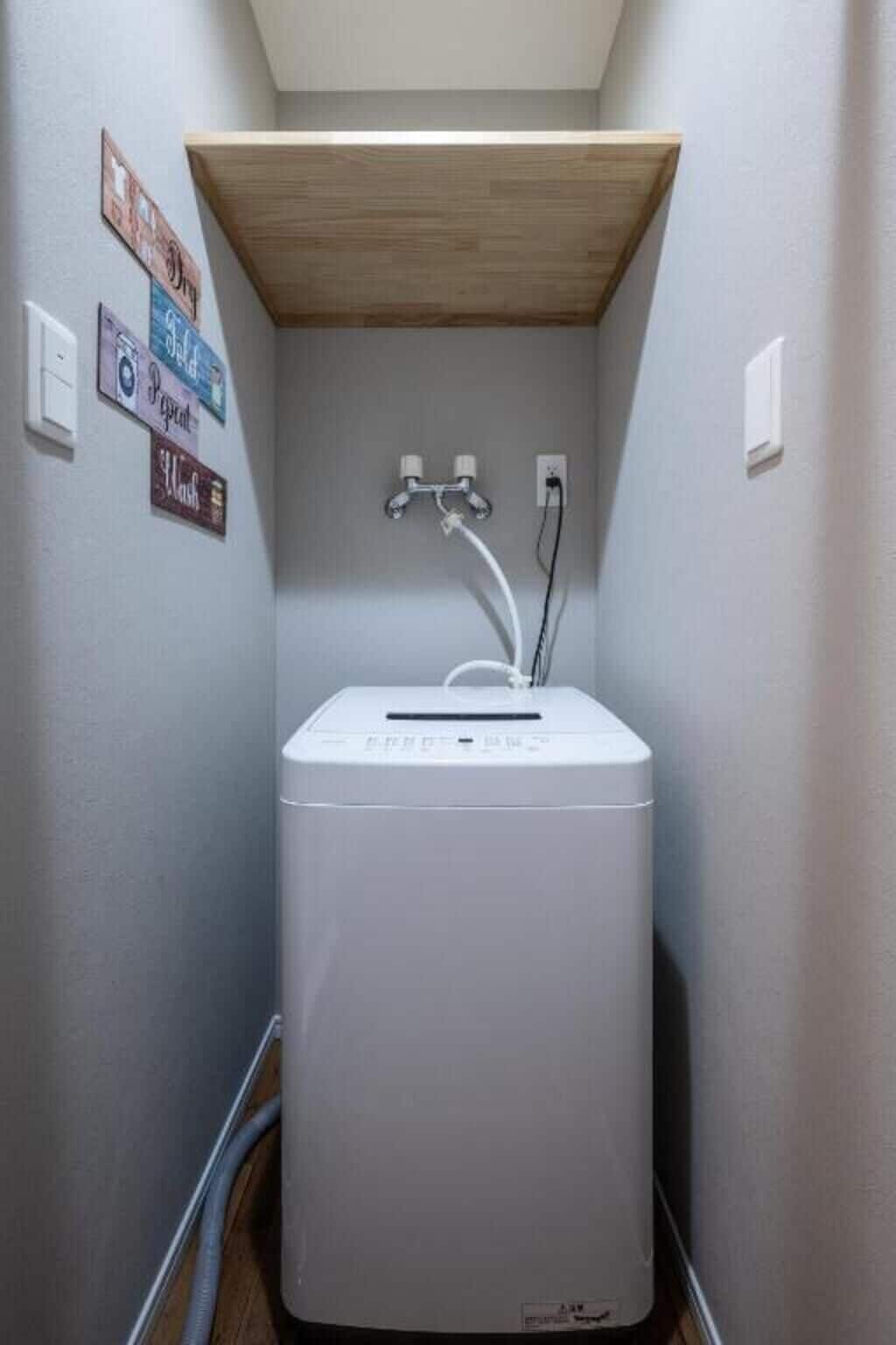 Lavadora blanca compacta en un espacio reducido, con un estante de madera encima y un enchufe eléctrico en la pared.