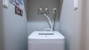 Lavadora blanca compacta en un espacio reducido, con un estante de madera encima y un enchufe eléctrico en la pared.