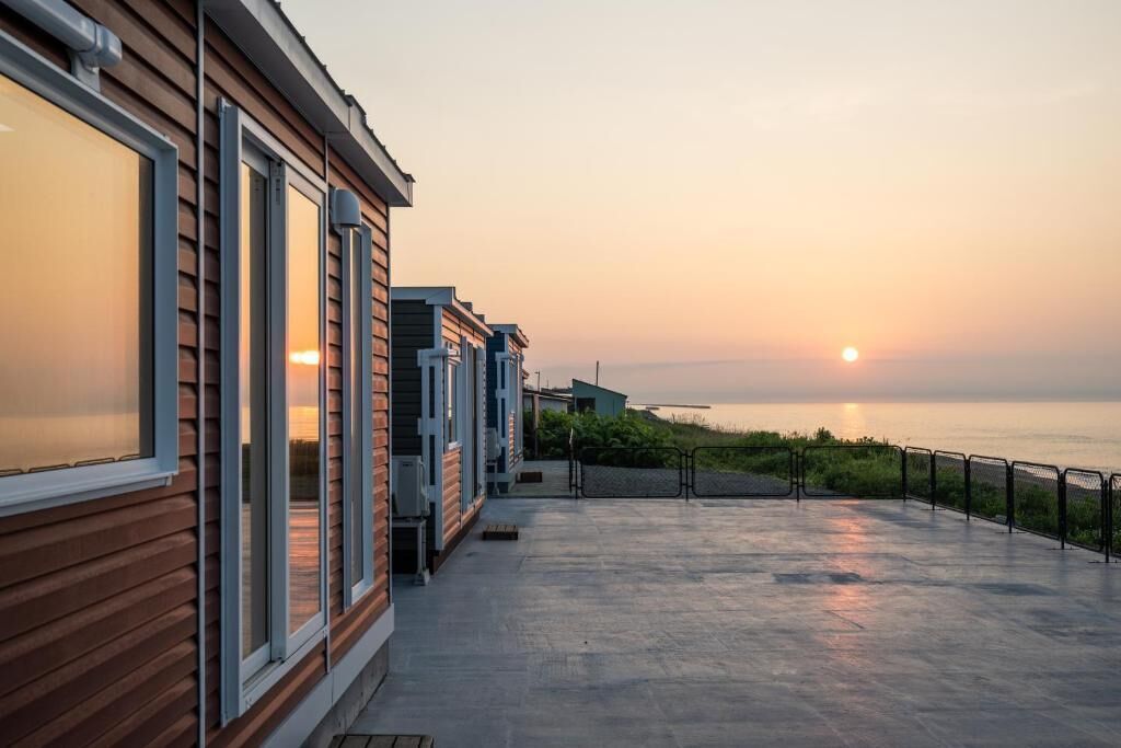 A row of wooden cabins with large windows, a concrete deck, and a view of the sea at sunset.