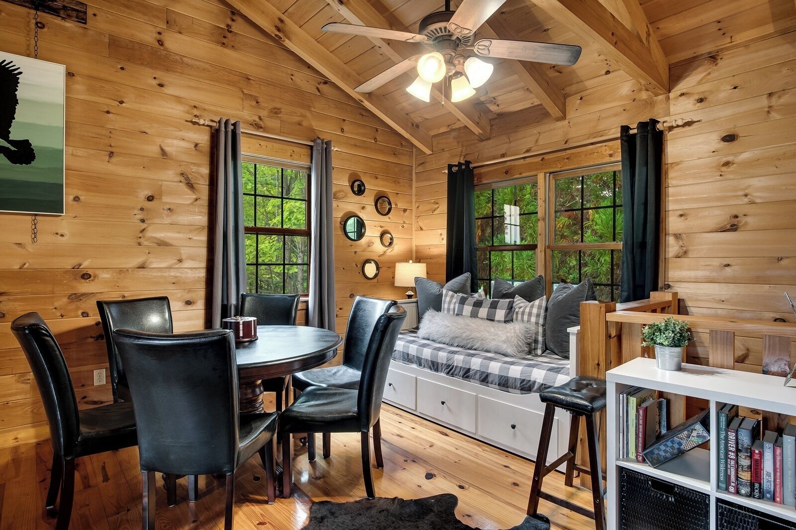 A cozy cabin interior with a dining area, a sofa with checkered cushions, and a bookshelf.