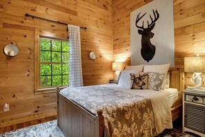 A wooden cabin bedroom with a bed, a window with curtains, a wall-mounted lamp, and a deer head artwork. - Cherokee Lookout Point (Bryson City)