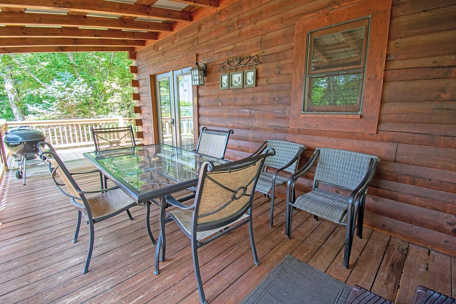 A wooden deck with a glass table and chairs, a barbecue grill, and a view of trees.