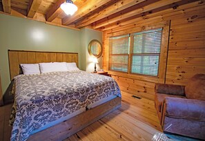 Ein Zimmer in einer Holzhütte mit einem Bett, einem Sessel, einem Fenster mit Jalousien und einer Deckenleuchte.