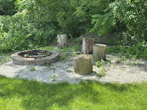 A garden with a circular stone fire pit surrounded by gravel, three wooden stumps, and dense foliage in the background.