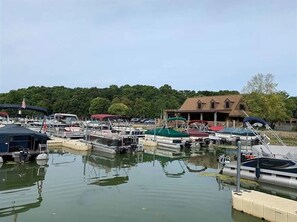 A marina with multiple boats docked, a building with a brown roof, and a forested area in the background.