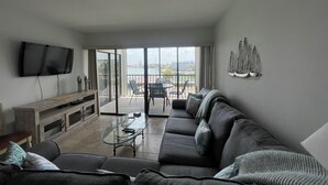 A living room with a sofa, a glass coffee table, a TV, and a balcony with a view. - Delightful and trendy 1 bedroom -1 bath condo F403 (Marco Island)
