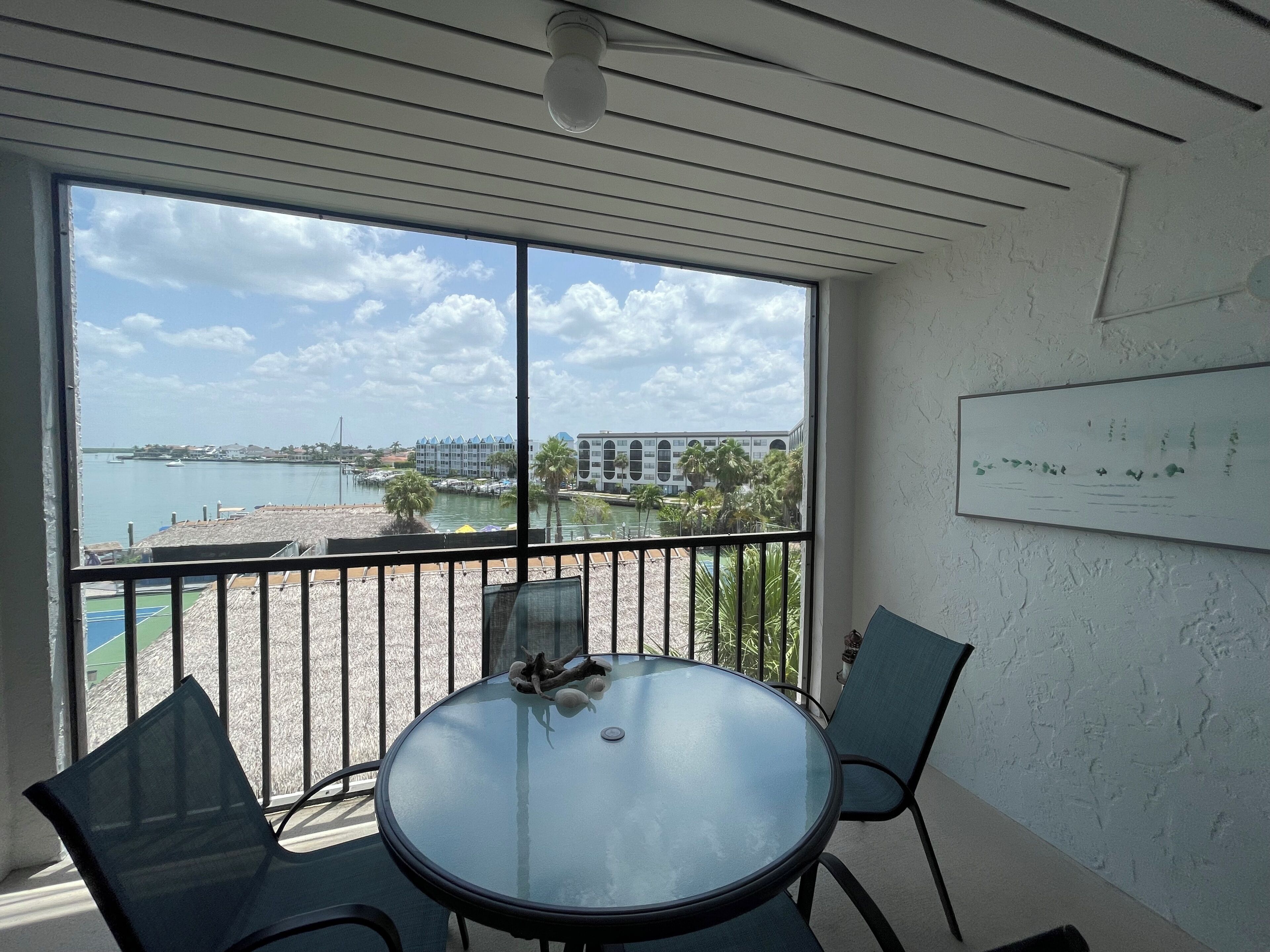 A room with a round glass table and two chairs, a view of a marina and buildings across the water, and a ceiling with a light fixture.