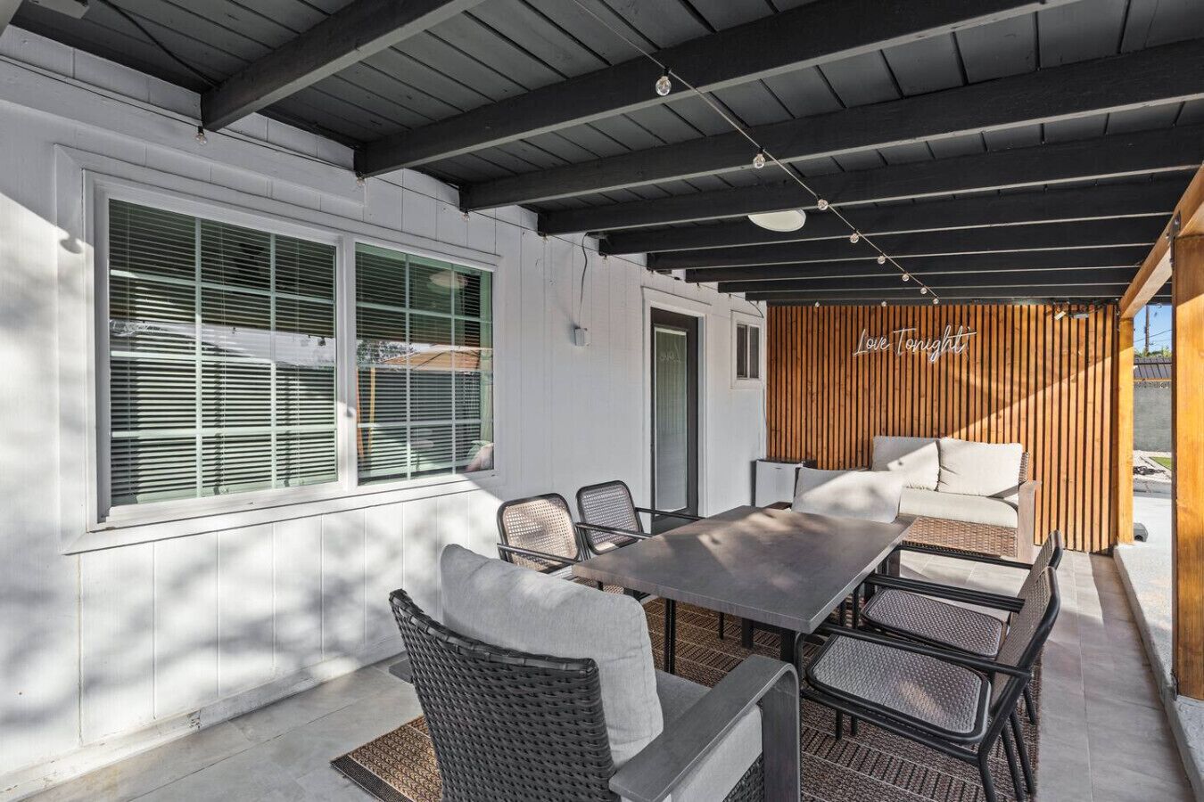 En udendørs terrasse med spisebord og stole, en trævæg med et skilt der siger 'Love Tonight', og et overdækket område med loft.