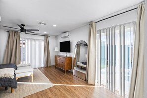 Ein modernes Hotelzimmer mit Holzboden, Flachbildfernseher, Deckenventilator, großem Spiegel und einem Bett mit Blick durch raumhohe Fenster.
