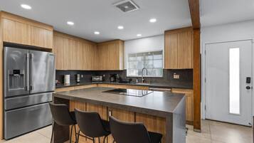 Uma cozinha moderna com frigorífico de aço inoxidável, armários de madeira, uma ilha central com banquetas e uma janela com persianas.