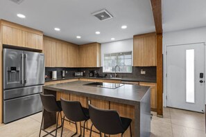 Eine moderne Küche mit einem Edelstahl-Kühlschrank, Holzschränken, einer Kochinsel mit Barhocker und einem Fenster mit Jalousien.