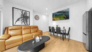 A modern living room with a leather sofa, a dining table with chairs, a black coffee table, a wall clock, and a flat-screen TV mounted above the dining area. - Richmond Escape | 5BR | 3 Baths | Walk to MCG (Cremorne)