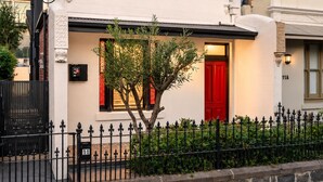 A white building with a red door, a black fence, and a tree in front. - Richmond Escape | 5BR | 3 Baths | Walk to MCG (Cremorne)