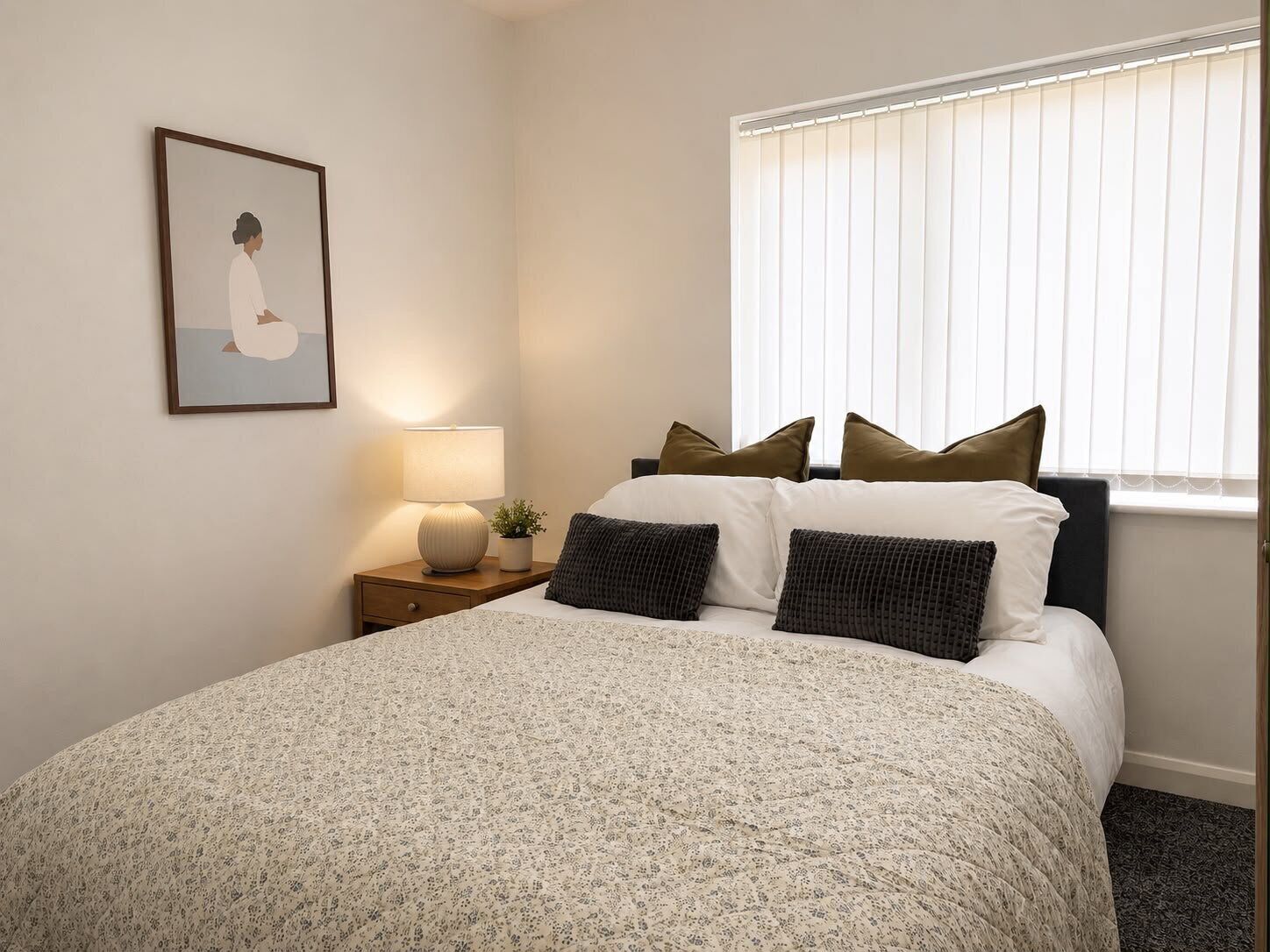 Ein Schlafzimmer mit einem Bett, Kissen, einem Nachttisch mit Lampe, einem gerahmten Bild an der Wand und einem Fenster mit Jalousien.