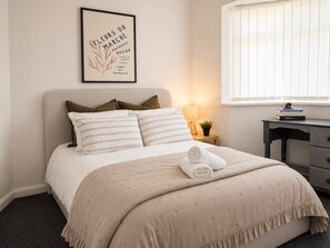 Un lit bien fait, recouvert d’une couvre-lit beige et agrémenté d’oreillers, un cadre décoratif accroché au mur, une table de chevet avec une lampe et une petite plante, et une fenêtre avec des stores.