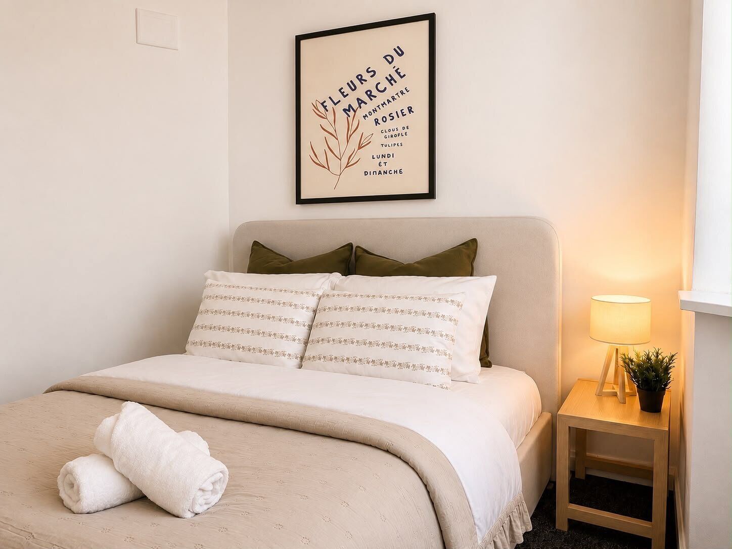 Un lit bien fait, avec une couette beige, deux oreillers et une serviette blanche roulée. Un cadre avec une citation y est accroché au mur, au-dessus du lit.