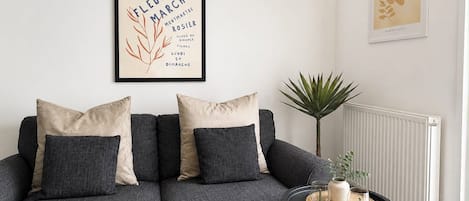 Un salon moderne avec un canapé gris, deux coussins, une œuvre d’art encadrée, une plante en pot et une petite table ronde sur laquelle se trouve un plateau.