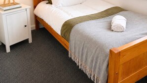 A single bed with a wooden frame, white bedding, a green throw blanket, and a rolled towel on the footboard. A bedside table with a lamp and a book is visible.