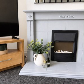 A fireplace with a white mantel, a television on a wooden stand to the left, and a vase with yellow flowers on the mantel. - Hartlepool 4BR Stay | Long Stay & Contractor Deals (Hartlepool)