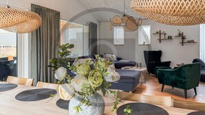 Een moderne woonkamer met een eethoek, met een stenen vaas met bloemen, geweven lampen en een donkerblauwe bank.
