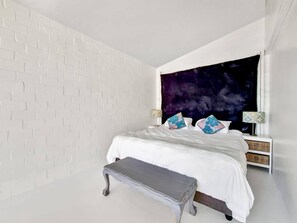 Ein Schlafzimmer mit weißer Ziegelwand, einem großen Bett mit weißer Bettwäsche und dekorativen Kissen, einer Bank und einem Nachttisch mit Lampen.