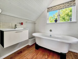 Ein Badezimmer mit einer freistehenden Badewanne, einem weißen Waschtisch mit Spiegel und einem Fenster mit Blick auf Grünflächen.