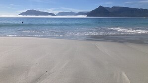 A beach with a person swimming, calm water, and distant mountains. - Kommetjie Luxury Retreat for Families & Groups (Cape Town)