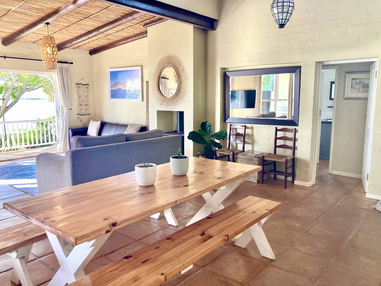 A wooden dining table with benches, a sofa, a large mirror, and a potted plant in a room with a tiled floor and a view of the outdoors.