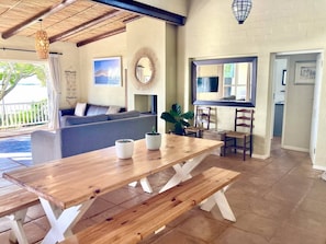 Ein Holztisch mit Bänken, eine Couch, ein großer Spiegel und ein Blumentopf in einem Raum mit Fliesenboden und Blick ins Freie.
