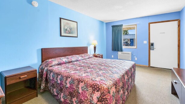 Habitación de hotel con cama, mesita de noche, lámpara, unidad de aire acondicionado y una puerta con el letrero 'No Smoking'.