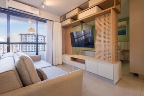 Ein modernes Wohnzimmer mit einer Couch, einem Fernseher auf einem Holzboden und Blick auf eine Stadtlandschaft durch große Fenster.