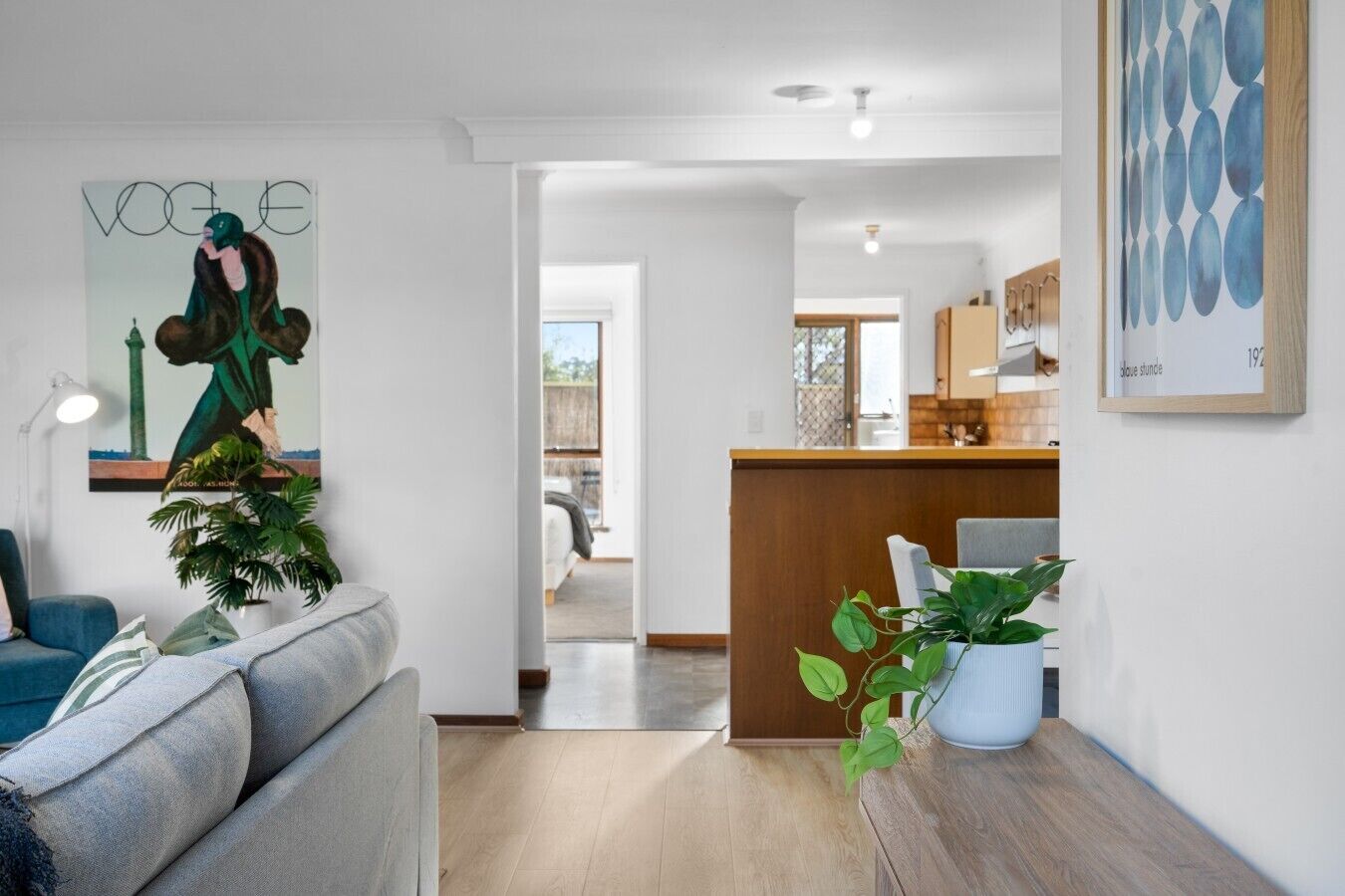 A modern living room with a sofa, a coffee table, and a potted plant. There is a kitchen area with wooden cabinets and a countertop visible through an open doorway.