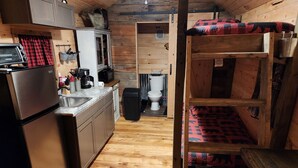 Cocina compacta con refrigerador, pileta y armarios de madera. Se ve una cama superpuesta con un edredón estampado al fondo.