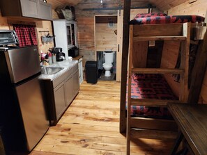 A compact kitchen with a refrigerator, sink, and wooden cabinets. A bunk bed with a patterned blanket is visible in the background. - Tranquil 1-bedroom cabin with AC in charming Zoe (Zoe)