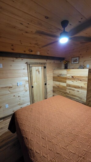 A room with wooden walls, a bed with a patterned bedspread, a ceiling fan, and a door. - Tranquil 1-bedroom cabin with AC in charming Zoe (Zoe)