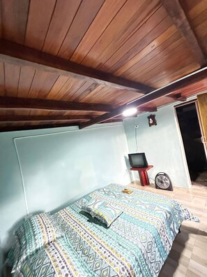 Un dormitorio con cama, televisor sobre un mueble, ventilador y techo de madera.