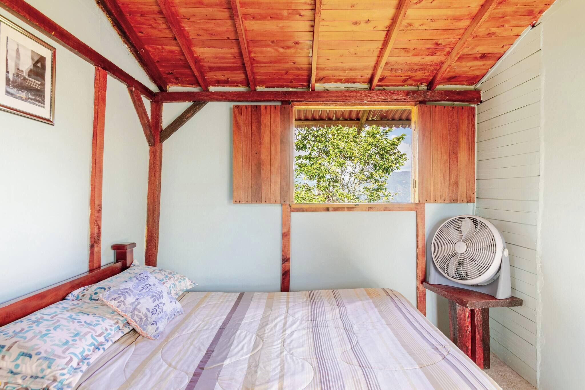 Una habitación con cama, techo de madera, ventana con vistas y un ventilador.