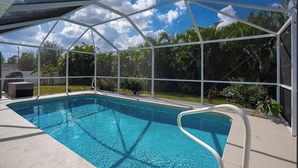 A clear blue pool with a white pool ladder, surrounded by a covered area with a white frame and a view of palm trees and a cloudy sky. - Private Heated Pool, Sleeps 10, King Suite, Near Dining and Boating (Cape Coral)