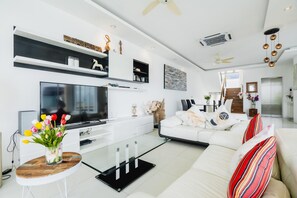 Ein modernes Wohnzimmer mit einem Flachbildfernseher, weißen Sofas, einem Glastisch und einer dekorativen Vase mit Blumen.