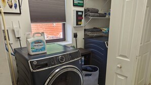 Ett kompakt tvättrum med en tvättmaskin och en torktumlare med frontmatning, ett fönster med persienner, en hylla med hopvikta handdukar och en tvättkorg.