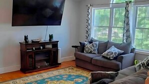 Sala de estar con televisor de pantalla plana, mueble de madera oscura para entretenimiento, sofá gris con cojines estampados, alfombra con diseño y una ventana con cortinas.