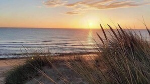 Une scène de plage avec le soleil se couchant sur l’océan, de l’herbe au premier plan et un ciel dégagé.