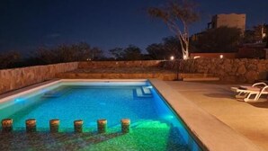 A swimming pool at night with a stone wall and a tree in the background.