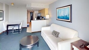 Ein Hotelzimmer mit weißem Sofa, rundem Couchtisch, Esstisch mit Stühlen, einer Küchenzeile und einem Segelboot-Gemälde an der Wand.