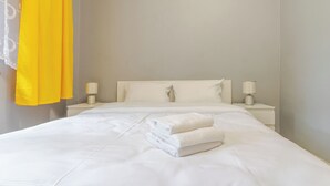 Un lit bien fait, avec du linge de lit blanc, deux lampes de chevet et un rideau jaune vif.