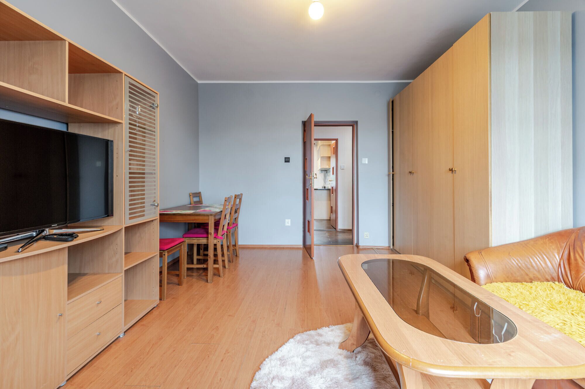 A modern living room with a flat-screen TV, wooden furniture, a dining area, and a walk-in closet.
